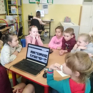 Nauczycielka i grupa  dzieci siedzą przy stolikach. Przed nauczycielką laptop z modelami 3d znanych polskich osób. Dzieci słuchają co mówi pani, uśmiechają się.