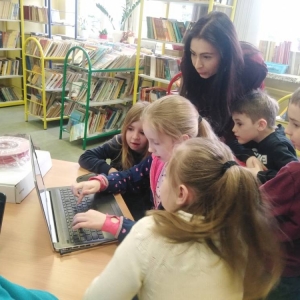 Dziewczynka siedzi przy laptopie i przegląda modele 3d online. Kilkoro dzieci i nauczycielka przyglądają się wyświetlanym przez nią modelom.