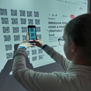 Uczennica, podczas zajęć j. angielskiego, przy pomocy smartfonu, wybiera i skanuje kod QR, spośród wyświetlonych na tablicy interaktywnej.