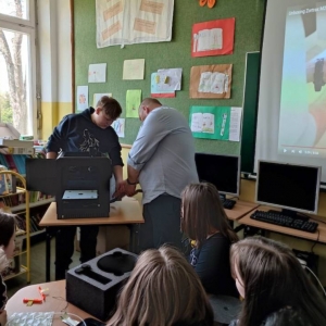 Nauczyciel i ósmoklasista podłączają drukarkę 3d zgodnie z instrukcją wyświetlaną na projektorze. Przy stoliku siedzi 5 ósmoklasistek.