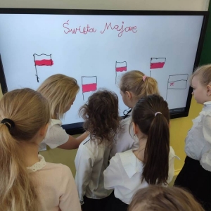 Grupa uczniów stoi przy tablicy interaktywnej. Nauczycielka tłumaczy im jej funkcjonalności.