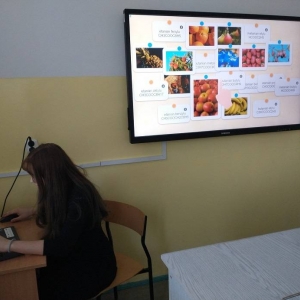Ósmoklasistka, podczas zajęć chemii, siedzi przy biurku przed laptopem, i rozwiązuje tematyczne zadania online, z wykorzystaniem aplikacji learningapps, wyświetlone również na ekranie telewizora za nią.