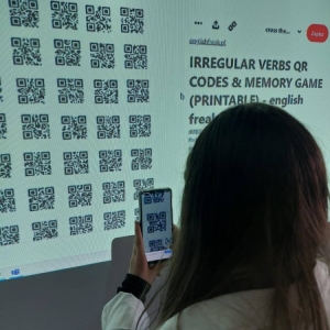 Szóstoklasistka, podczas zajęć j. angielskiego, przy pomocy smartfonu, wybiera i skanuje kod QR, spośród wyświetlonych na tablicy interaktywnej.