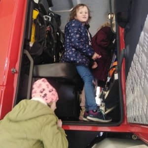 Dwie dziewczynki stoją w środku kabiny wozu strażackiego. Kolejna wchodzi do środka po schodkach.