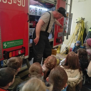 Przedstawiciel OSP pokazuje i opisuje dzieciom sprzęty znajdujące się na tyle wozu strażackiego. Dzieci stoją, przyglądają się i słuchają.