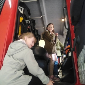 Jedna uczennica stoi w kabinie wozu strażackiego, druga wchodzi do środka.