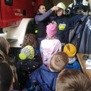 Przedstawiciele OSP wzajemnie pomagają sobie ubrać stój strażacki. Dzieci stoją i się przyglądają.