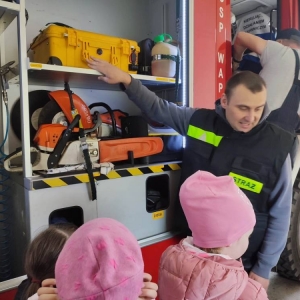 Przedstawiciel OSP pokazuje i opisuje dzieciom sprzęty znajdujące się w bocznej części wozu strażackiego.