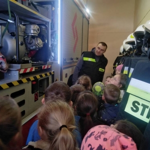 Przedstawiciel OSP o liczna grupa dzieci stoją z boku wozu strażackiego i oglądają jego wnętrze.