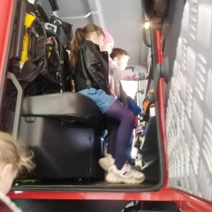 Dwie dziewczynki i chłopiec stoją w środku kabiny wozu strażackiego. Kolejna wchodzi do środka po schodkach.