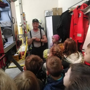 Przedstawiciel OSP o liczna grupa dzieci stoją na tyle wozu strażackiego i oglądają jego wnętrze.
