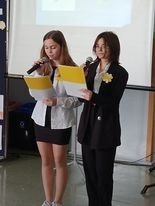 Dwie dziewczynki ubrane na galowo na tle tablicy z prezentacją prowadzą apel poświęcony patronowi szkoły. 